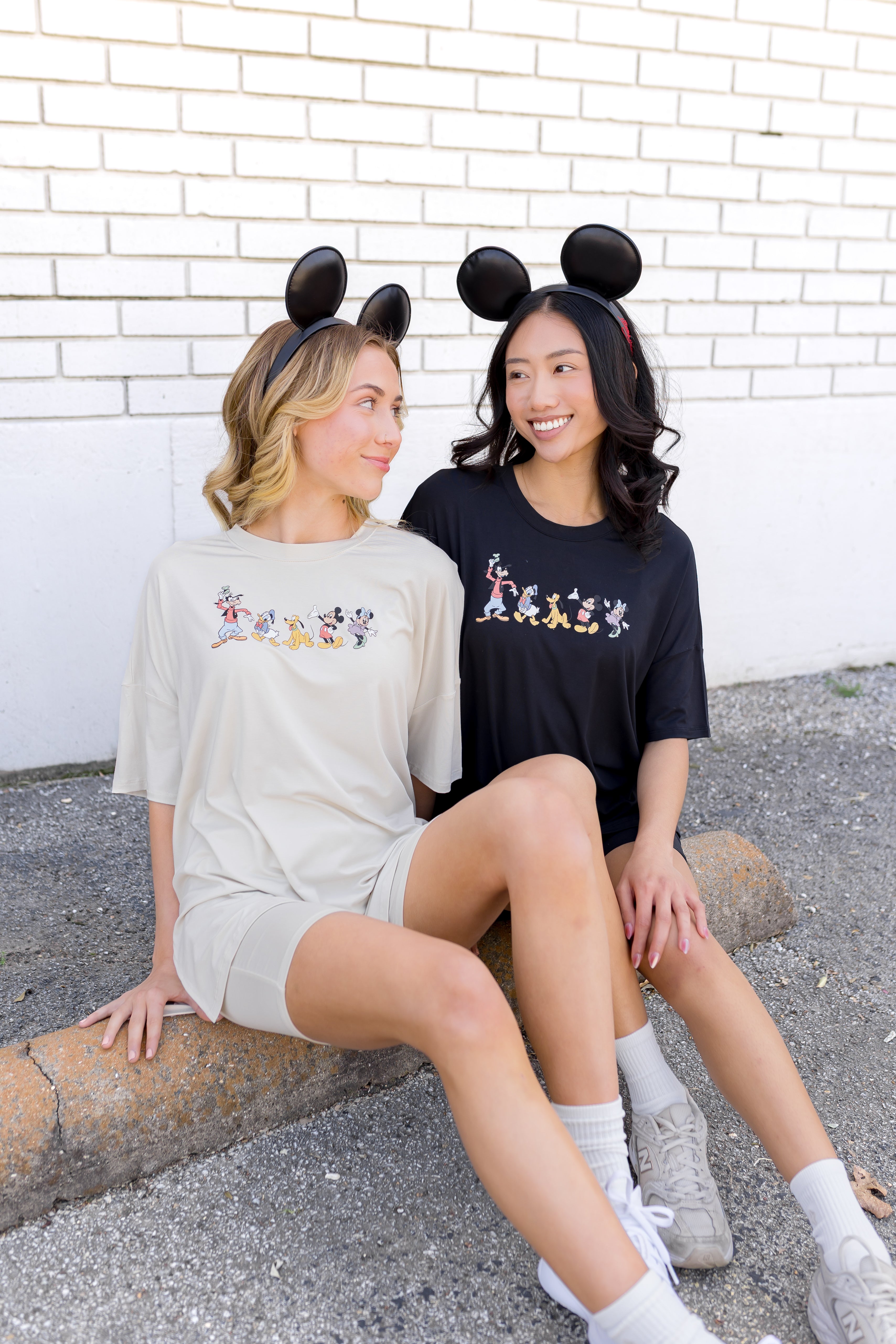 2 women wearing biker short sets in a cream and black colorway with mickey and friend graphic and wearing mickey mouse ear headbands