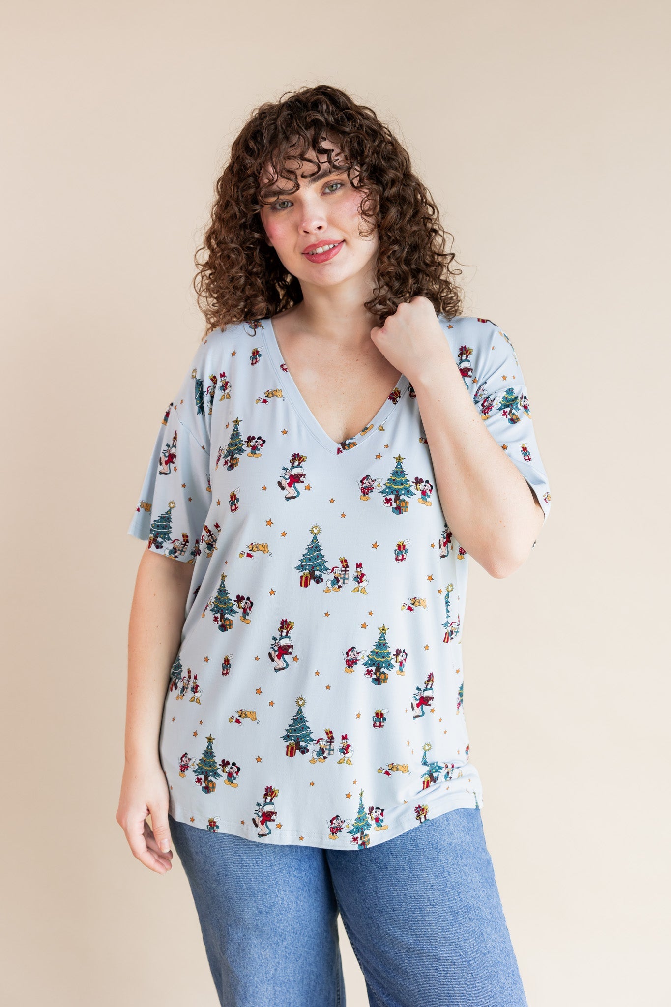 Woman wearing Women's Oversized V-Neck Tee in Mickey & Friends Christmas print over blue jeans on a beige background

