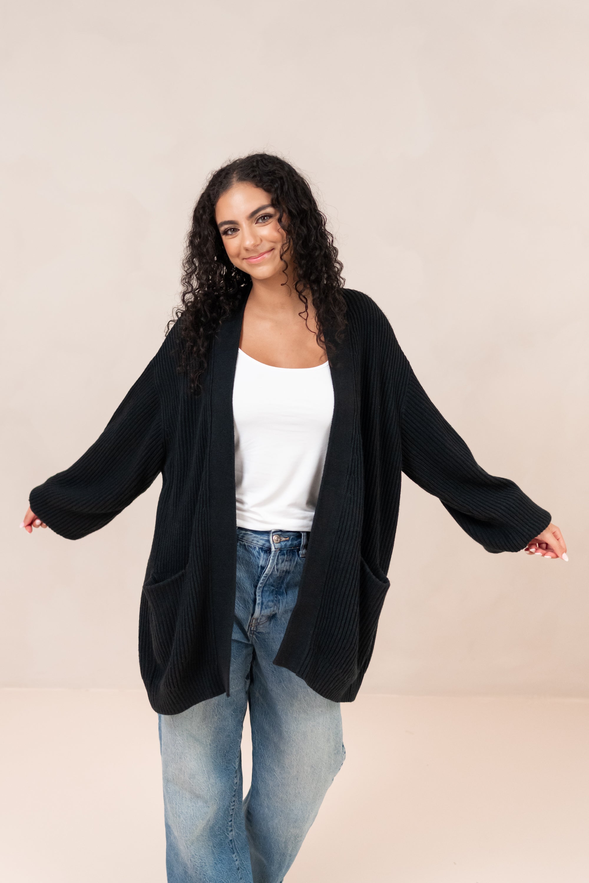 Woman wearing a black cardigan over a white top and blue jeans on a beige background