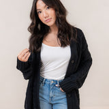 Woman wearing a black cardigan, white top, and blue jeans against a plain background
