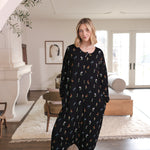 Model standing in a light modern bohemian room while wearing the Long Sleeve Adult Sleep Bag Walker in Midnight Wildflower with her hands in her pocket ::: Model is 5'10" wearing size XS ///