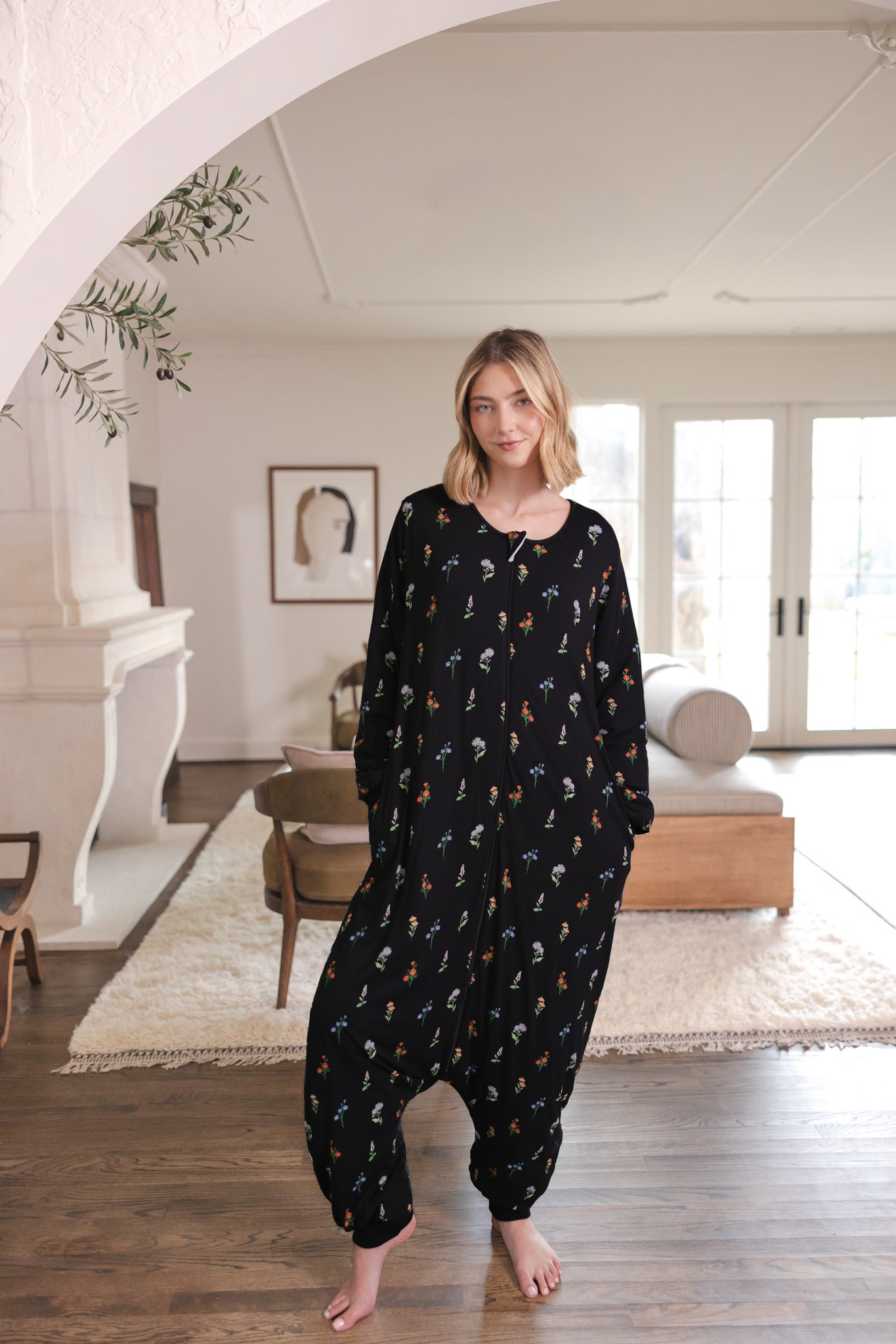 Model standing in a light modern bohemian room while wearing the Long Sleeve Adult Sleep Bag Walker in Midnight Wildflower with her hands in her pocket 