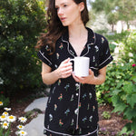 Close up of a model holding a white mug and modeling the Women's Short Sleeve Pajama Set in Midnight Wildflower in a garden ---PJ 4