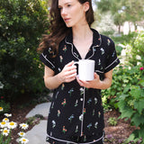 Close up of a model holding a white mug and modeling the Women's Short Sleeve Pajama Set in Midnight Wildflower in a garden ---PJ 4