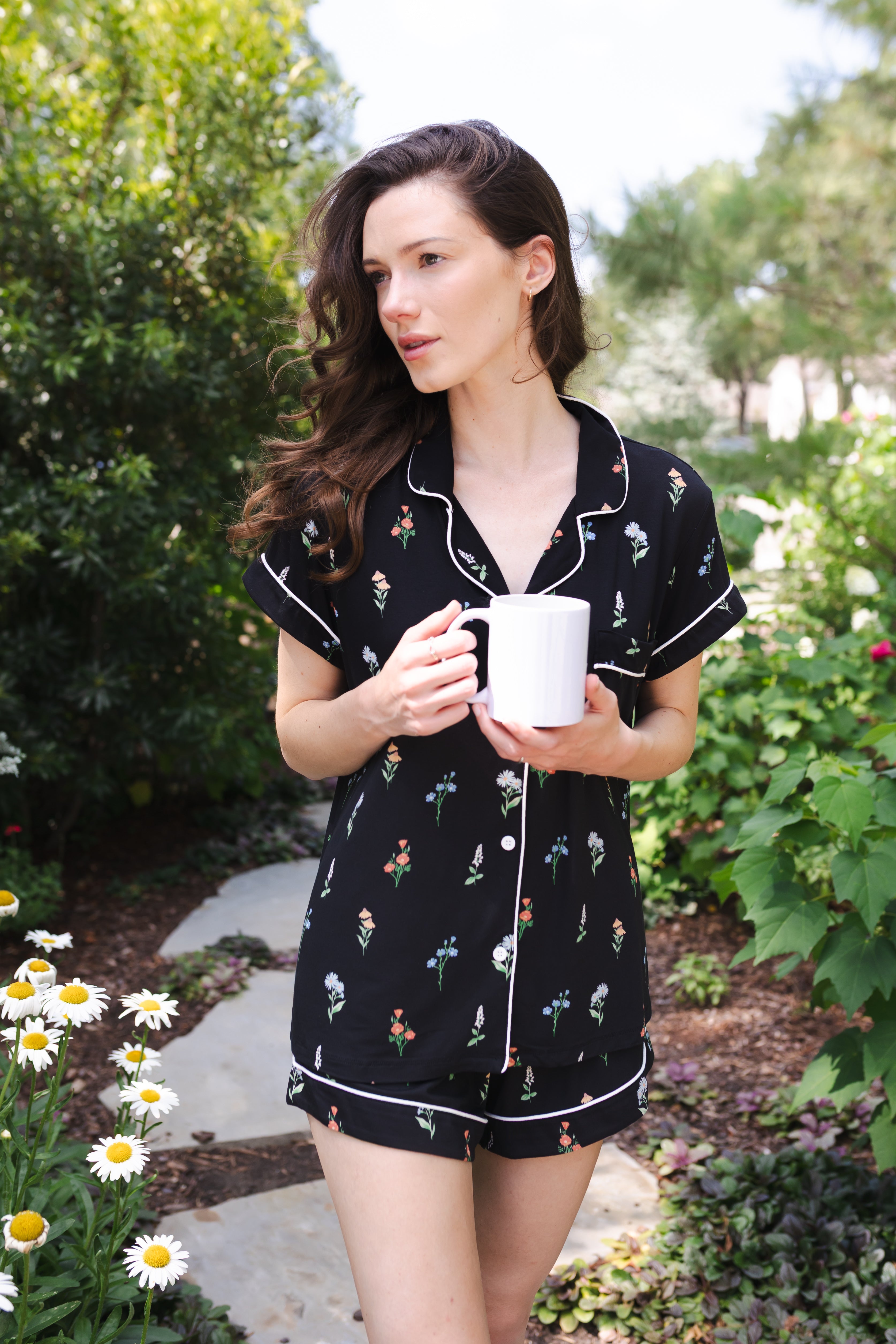 Close up of a model holding a white mug and modeling the Women's Short Sleeve Pajama Set in Midnight Wildflower in a garden ---PJ 4