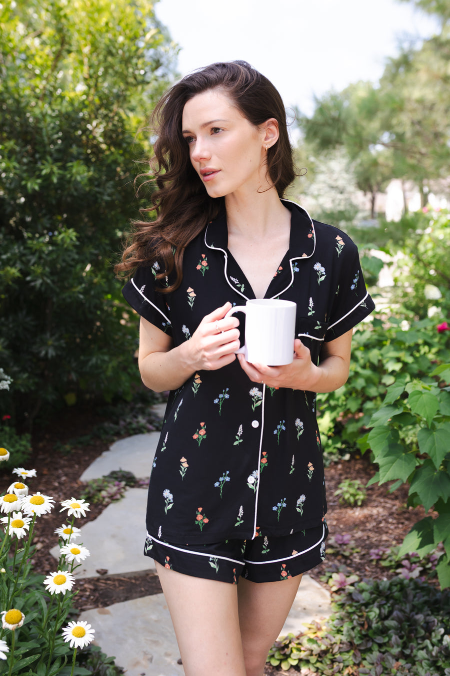 Close up of a model holding a white mug and modeling the Women's Short Sleeve Pajama Set in Midnight Wildflower in a garden