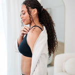 Woman wearing a black cross front bralette and midi brief with oat robe in a bright room.