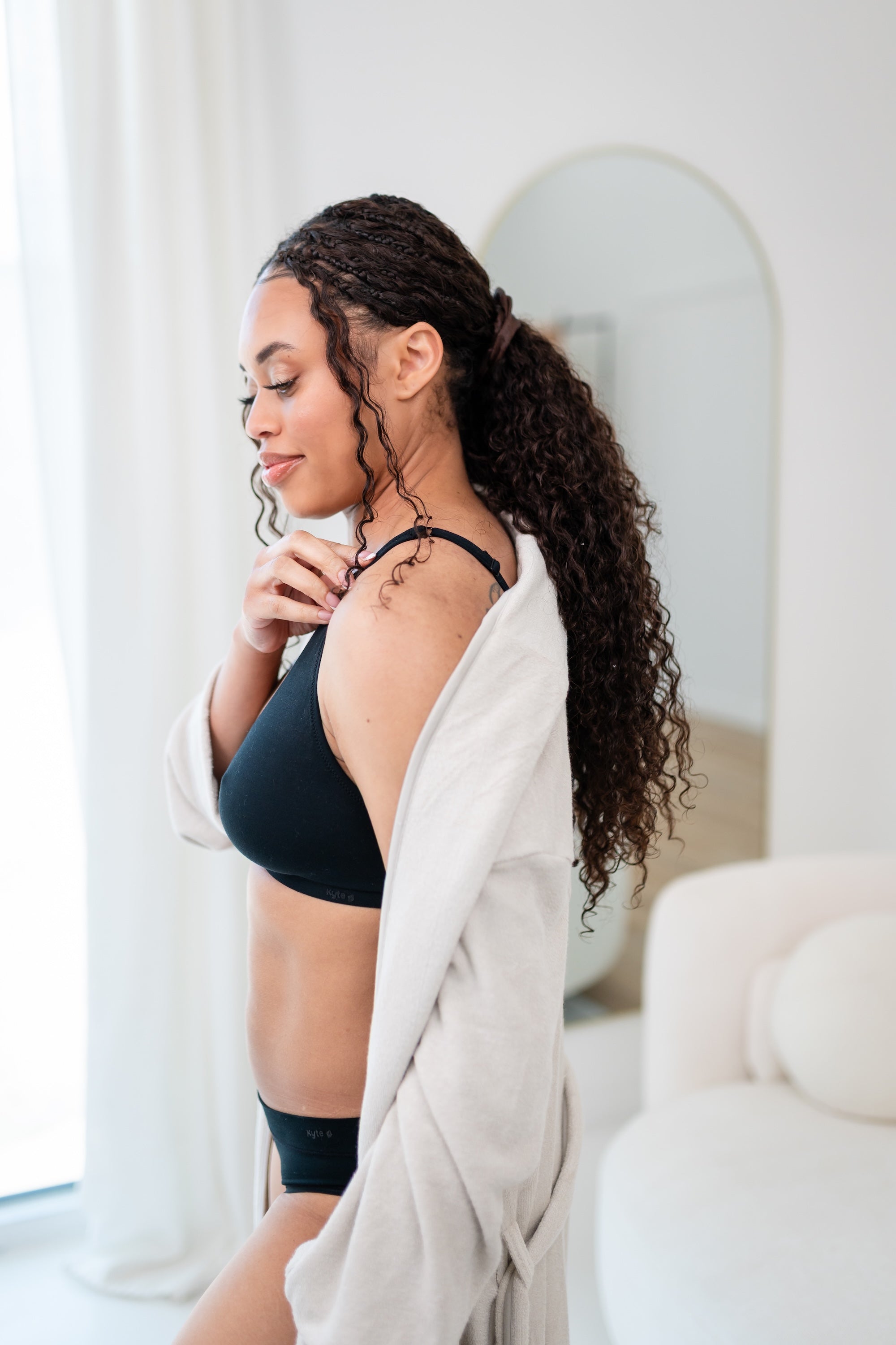 Woman wearing a black cross front bralette and midi brief with oat robe in a bright room.