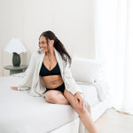 Woman in black crossfront bralette and midi brief sitting on a bed in a bright room with oat robe
