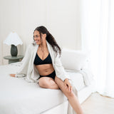 Woman in black crossfront bralette and midi brief sitting on a bed in a bright room with oat robe