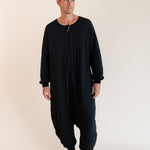 A man wearing a black long-sleeve, one-piece harem-style sleep sack  front zipper, standing barefoot against a plain beige studio background.