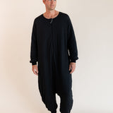 A man wearing a black long-sleeve, one-piece harem-style sleep sack  front zipper, standing barefoot against a plain beige studio background.