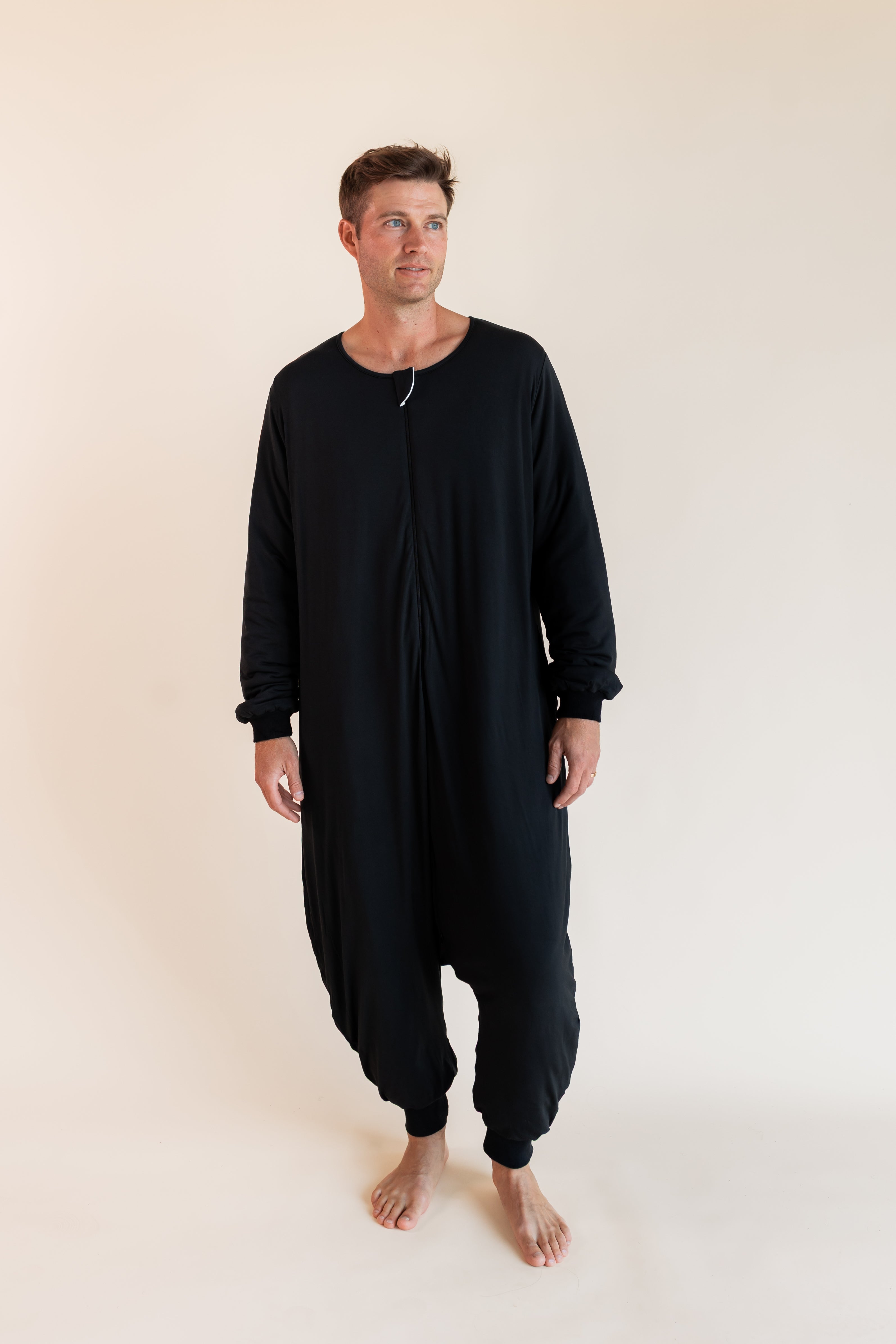A man wearing a black long-sleeve, one-piece harem-style sleep sack  front zipper, standing barefoot against a plain beige studio background.