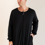 A man wearing a black long-sleeve sleep sack with a front zipper and side pockets, standing against a plain light beige studio background.