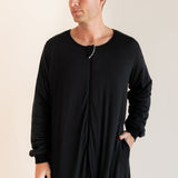 A man wearing a black long-sleeve sleep sack with a front zipper and side pockets, standing against a plain light beige studio background.
