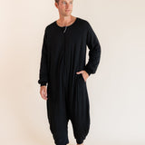 A man modeling a black long-sleeve harem-style sleep sack against a plain beige studio background.