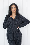 Woman wearing black button up pajamas in a monochromatic leopard print with white wooden wall backdrop