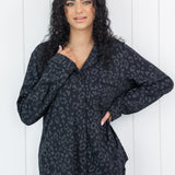 Woman wearing black button up pajamas in a monochromatic leopard print with white wooden wall backdrop