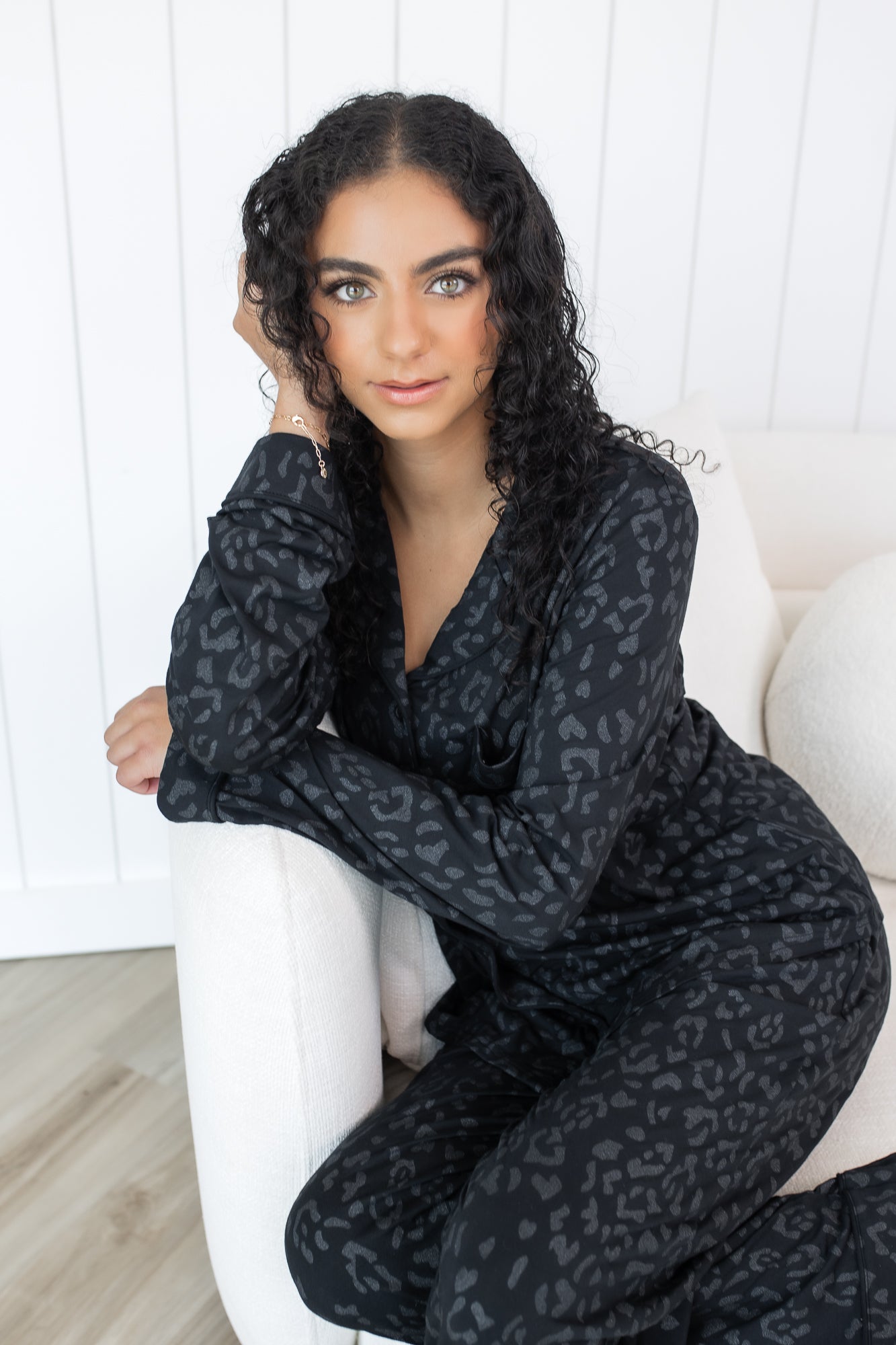Woman sitting in cream chair wearing black button up pajamas in a monochromatic leopard print