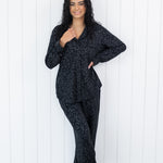Woman wearing black button up pajamas in a monochromatic leopard print against a white wooden wall backdrop