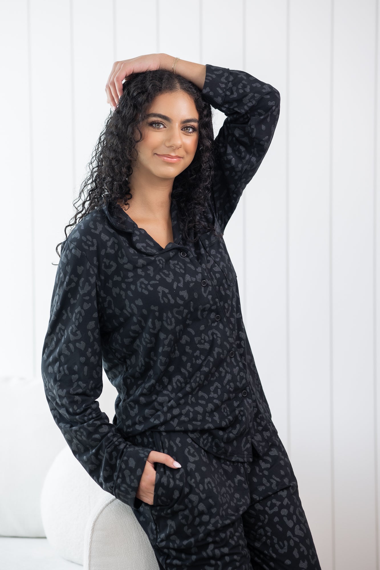 Woman leaning against a cream chair wearing black button up pajamas in a monochromatic leopard print