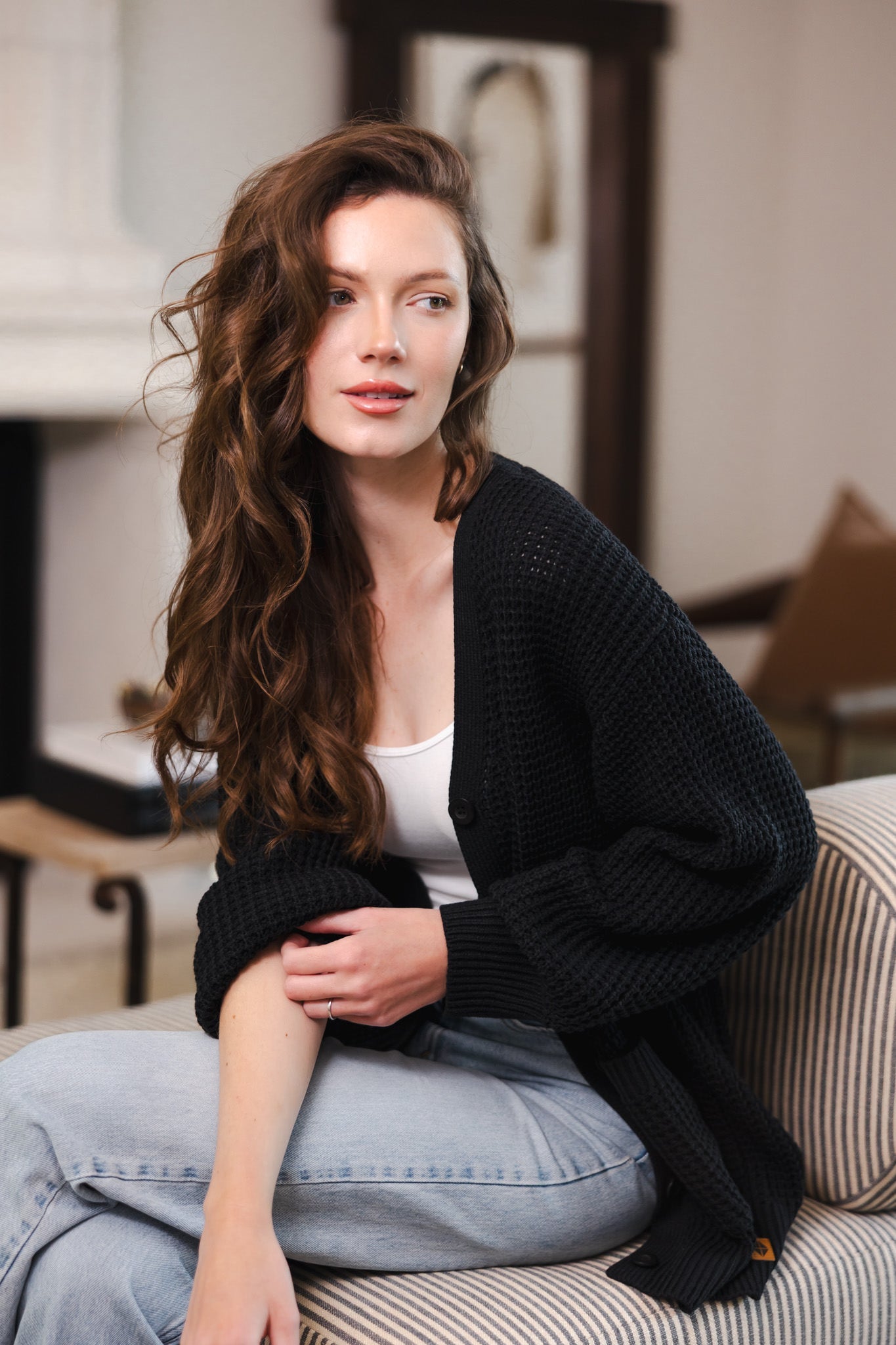 Modal sitting on a sofa in a living room wearing Chunky Knit Women's Waffle Cardigan in Midnight over white camisole and blue jeans
