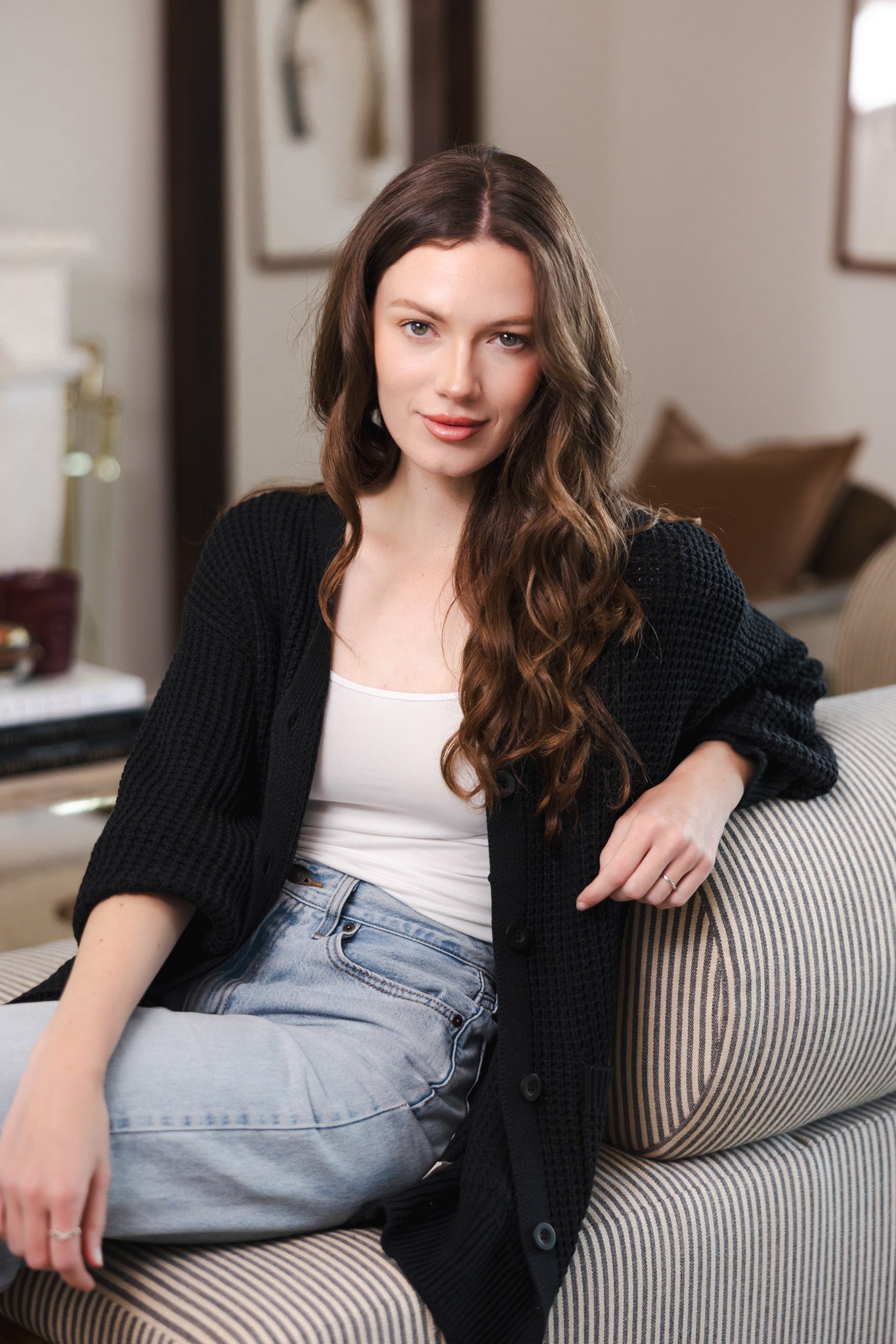 Modal posing on a sofa in a living room wearing Chunky Knit Women's Waffle Cardigan in Midnight over white camisole and blue jeans