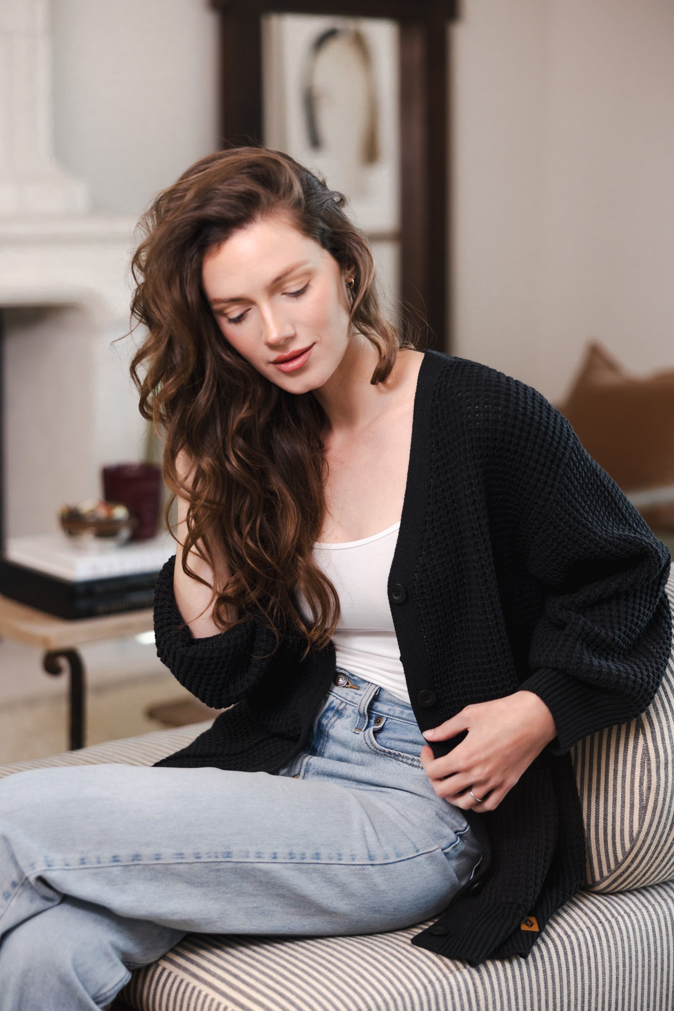 Modal sitting on a sofa in a living room wearing Chunky Knit Women's Waffle Cardigan in Midnight over white camisole and blue jeans