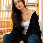 Woman sitting on a couch wearing a black cardigan, white top, and blue jeans in a room with wooden shelves.