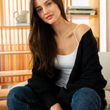 Woman sitting on a couch wearing a black cardigan, white top, and blue jeans in a room with wooden shelves.