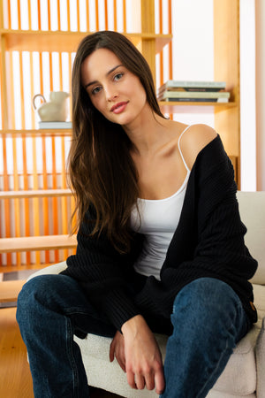 Woman sitting on a couch wearing a black cardigan, white top, and blue jeans in a room with wooden shelves.