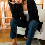 Woman sitting on a white couch wearing a black cardigan, white top, and blue jeans in a living room.