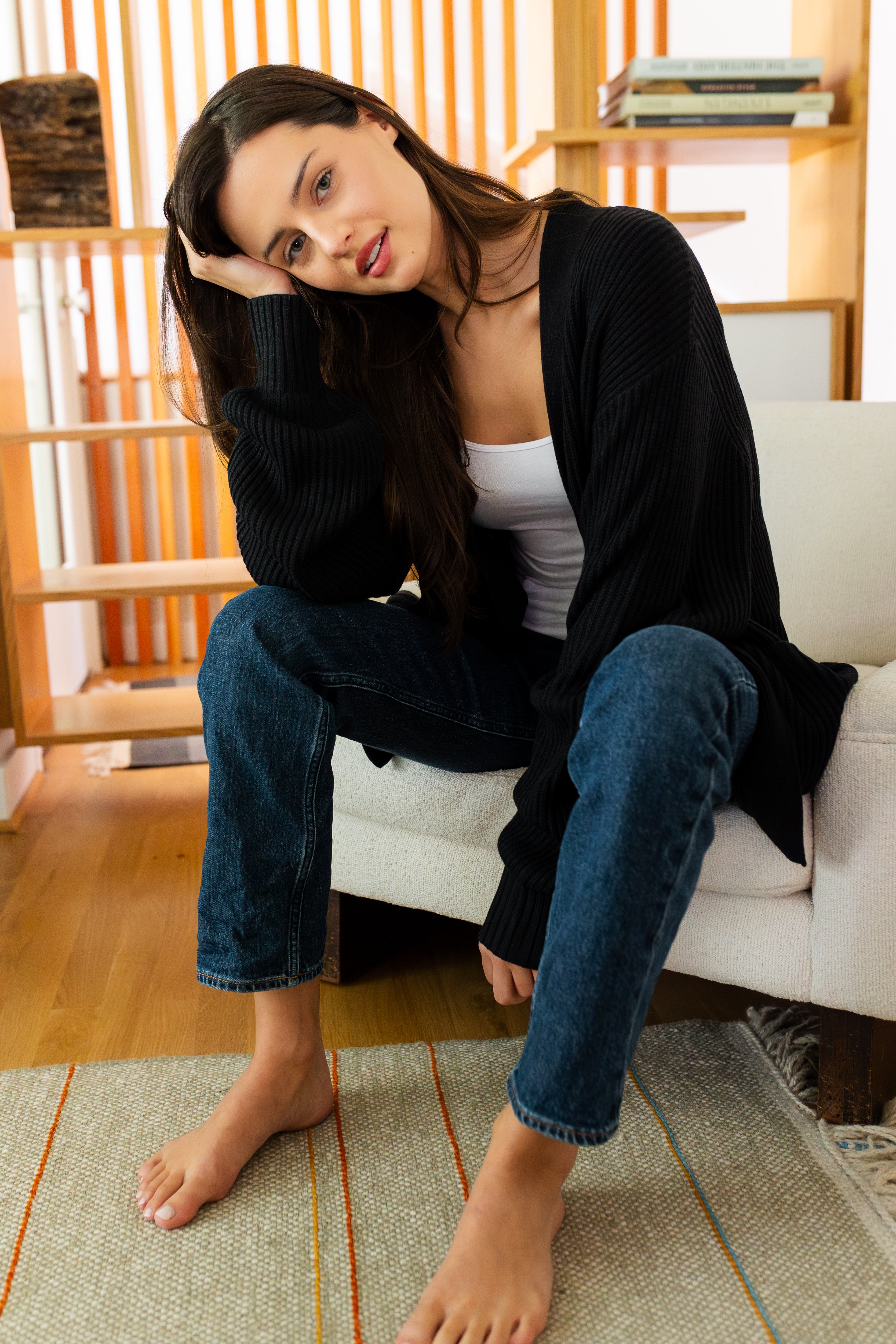 Woman sitting on a white couch wearing a black cardigan, white top, and blue jeans in a living room.