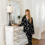 Woman in black magnolia floral pajamas standing in a room with a white dresser and mirror.