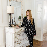 Woman in black magnolia floral pajamas standing in a room with a white dresser and mirror.
