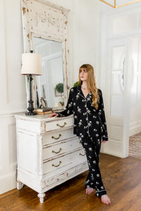 Woman in black magnolia floral pajamas standing in a room with a white dresser and mirror.