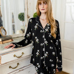 Woman wearing a black magnolia floral pajama set in a room with white walls and wooden floor. ::: Model is 5'6" wearing size S ///