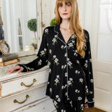 Woman wearing a black magnolia floral pajama set in a room with white walls and wooden floor. ::: Model is 5'6" wearing size S ///