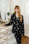 Woman wearing a black magnolia floral pajama set in a room with white walls and wooden floor. ::: Model is 5'6" wearing size S ///