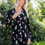 A woman wearing a black long-sleeve pajama set with a white floral print, standing outdoors in front of green foliage.