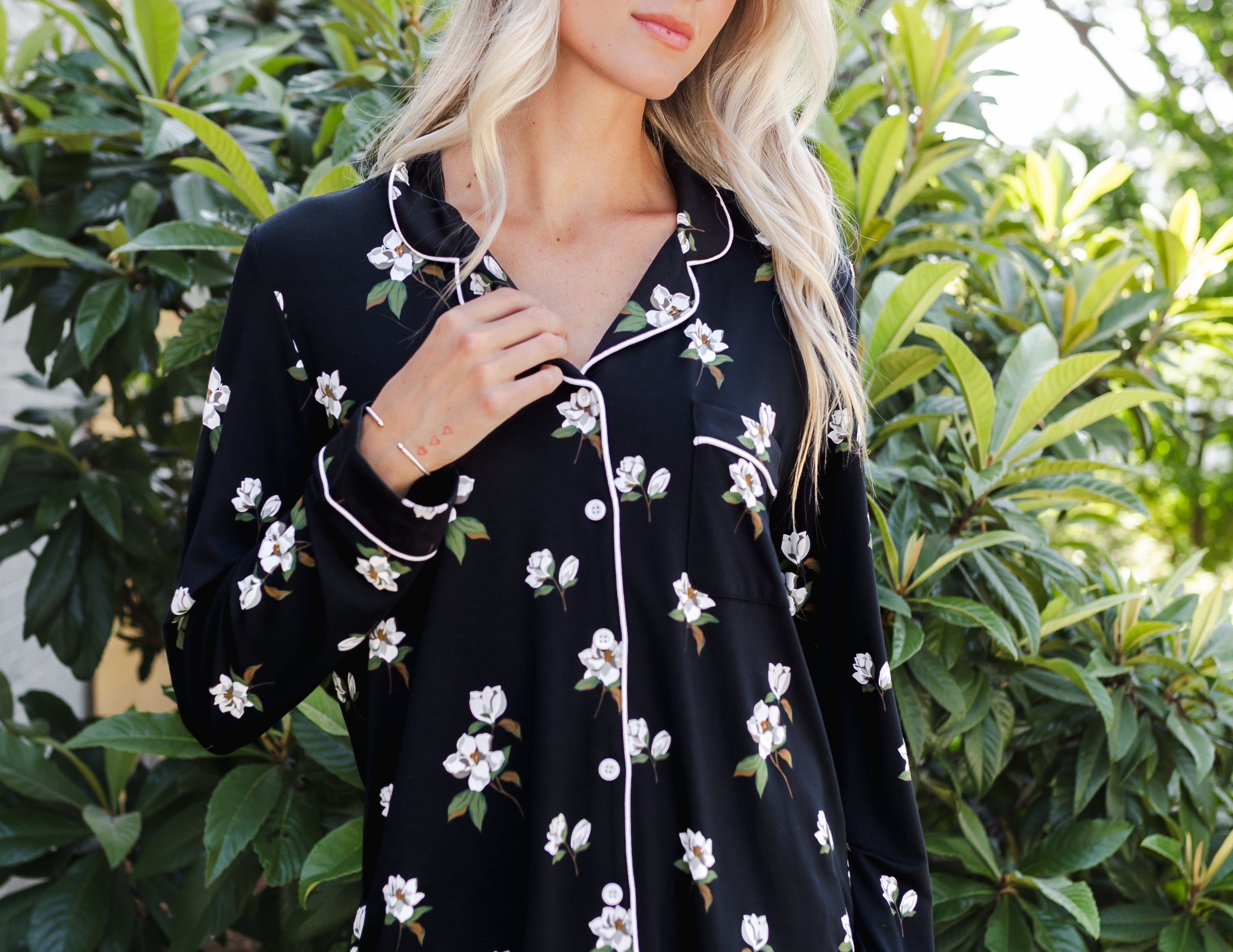 A woman wearing a black long-sleeve pajama shirt with white floral prints and white piping, standing outdoors in front of green foliage.
