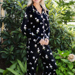 A woman wearing a black long-sleeve pajama set with a white floral print and white piping trim, standing outdoors in front of lush green garden foliage.