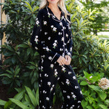 A woman wearing a black long-sleeve pajama set with a white floral print and white piping trim, standing outdoors in front of lush green garden foliage.