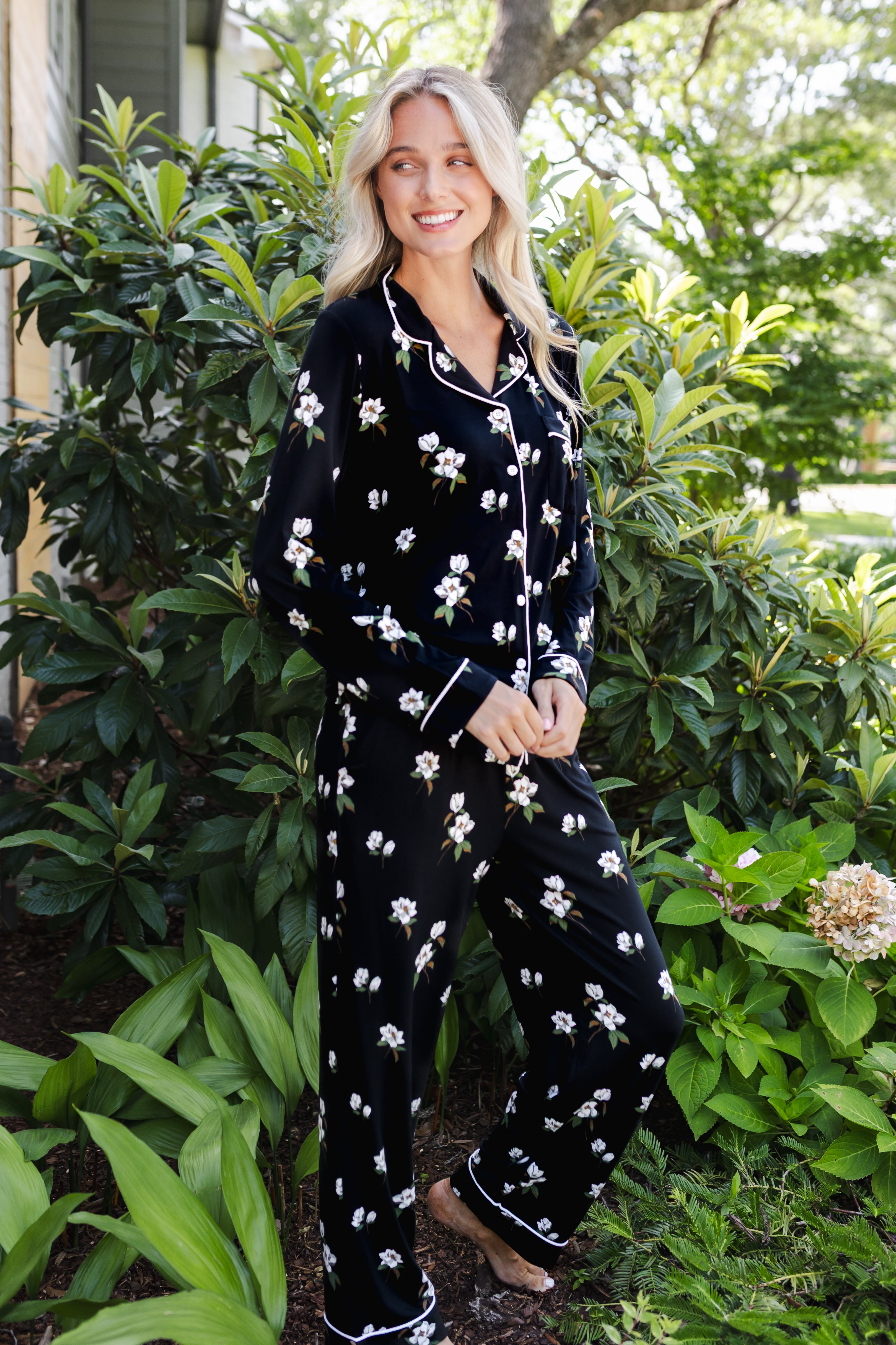 A woman wearing a black long-sleeve pajama set with a white floral print and white piping trim, standing outdoors in front of lush green garden foliage.