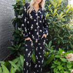 A woman wearing a black long-sleeve pajama set with a white floral print stands outdoors in front of green plants and a gray brick wall.