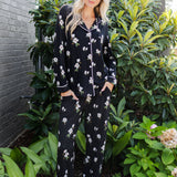 A woman wearing a black long-sleeve pajama set with a white floral print stands outdoors in front of green plants and a gray brick wall.