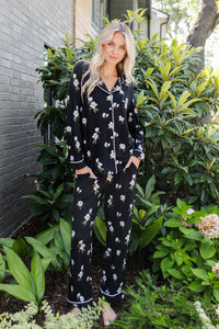 A woman wearing a black long-sleeve pajama set with a white floral print stands outdoors in front of green plants and a gray brick wall.