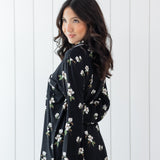 Woman modeling black long sleeve button up pajamas in a magnolia print against a white wooden wall