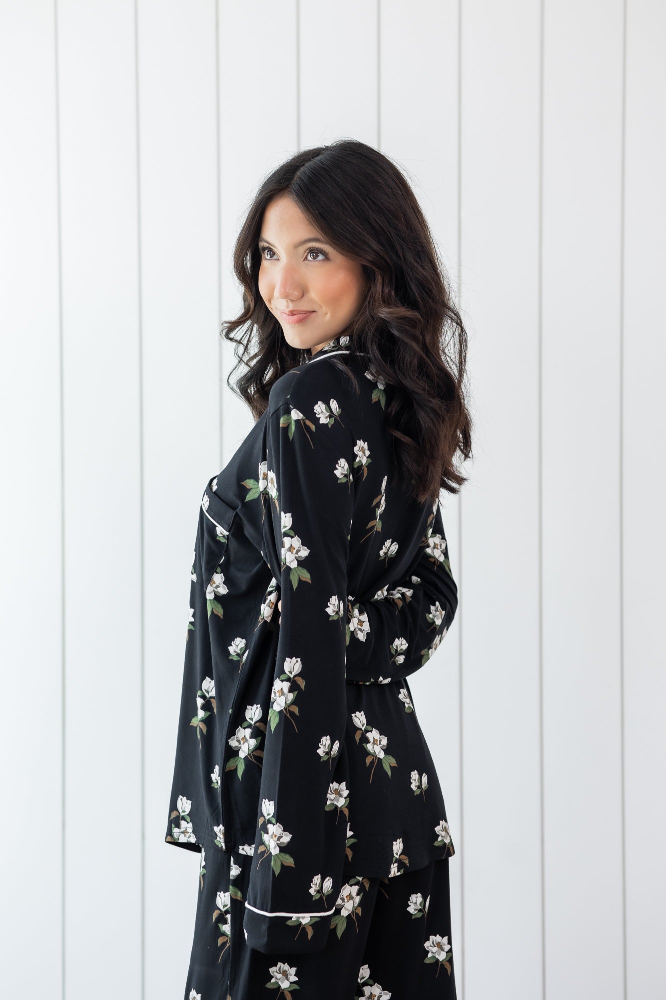 Woman modeling black long sleeve button up pajamas in a magnolia print against a white wooden wall