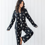 Woman modeling black long sleeve button up pajamas in a magnolia print against a white wooden wall